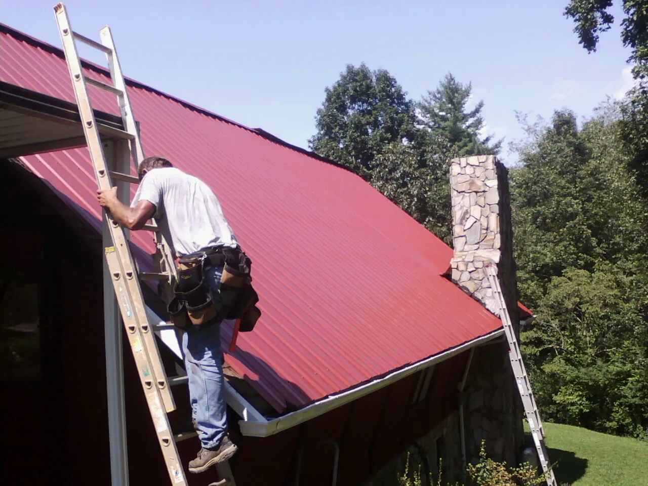Roofer on a ladder beside a red metal roof and stone chimney — Soffit & Fascia Repair by Roofing Brothers in Moonachie